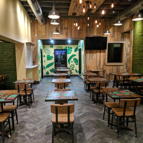 Empty restaurant dining room with wooden tables and chairs, wood-paneled walls, and tropical leaf murals.