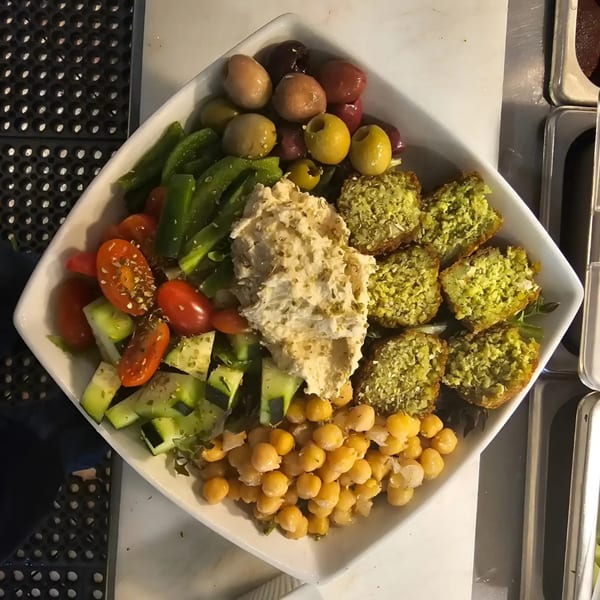 A square white bowl filled with falafel, hummus, chickpeas, olives, tomatoes, and cucumbers.