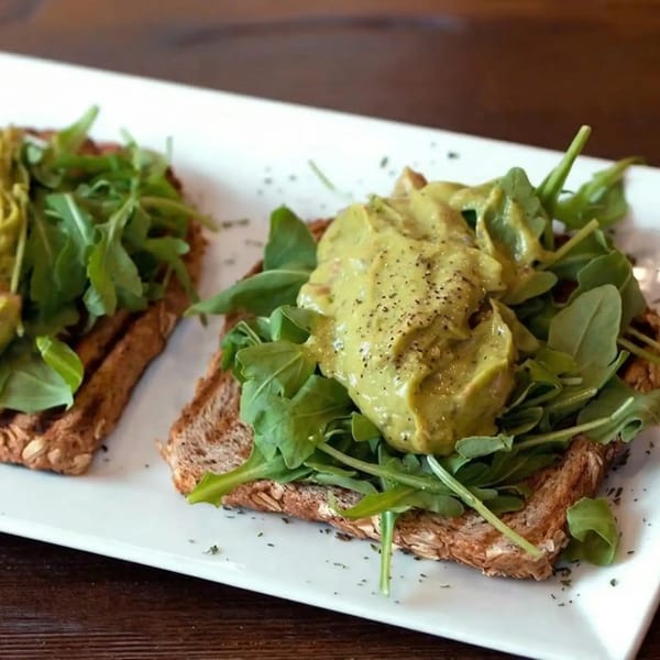 Two slices of avocado toast with arugula and pepper on a white rectangular plate.