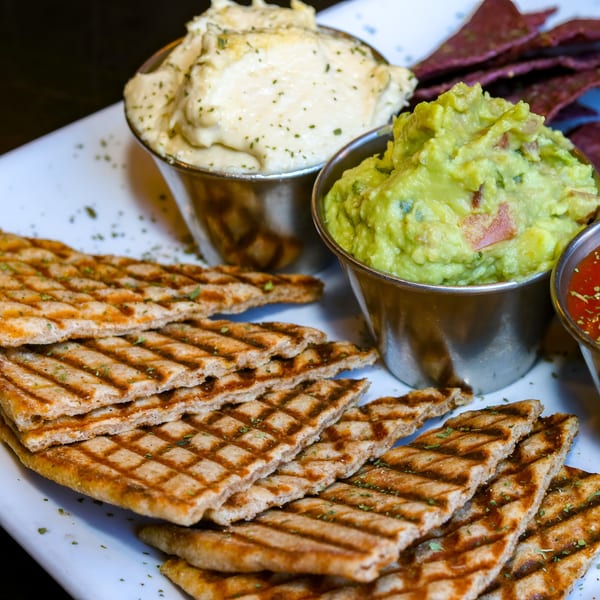 A platter with grilled flatbread, hummus, guacamole, salsa, and purple tortilla chips.