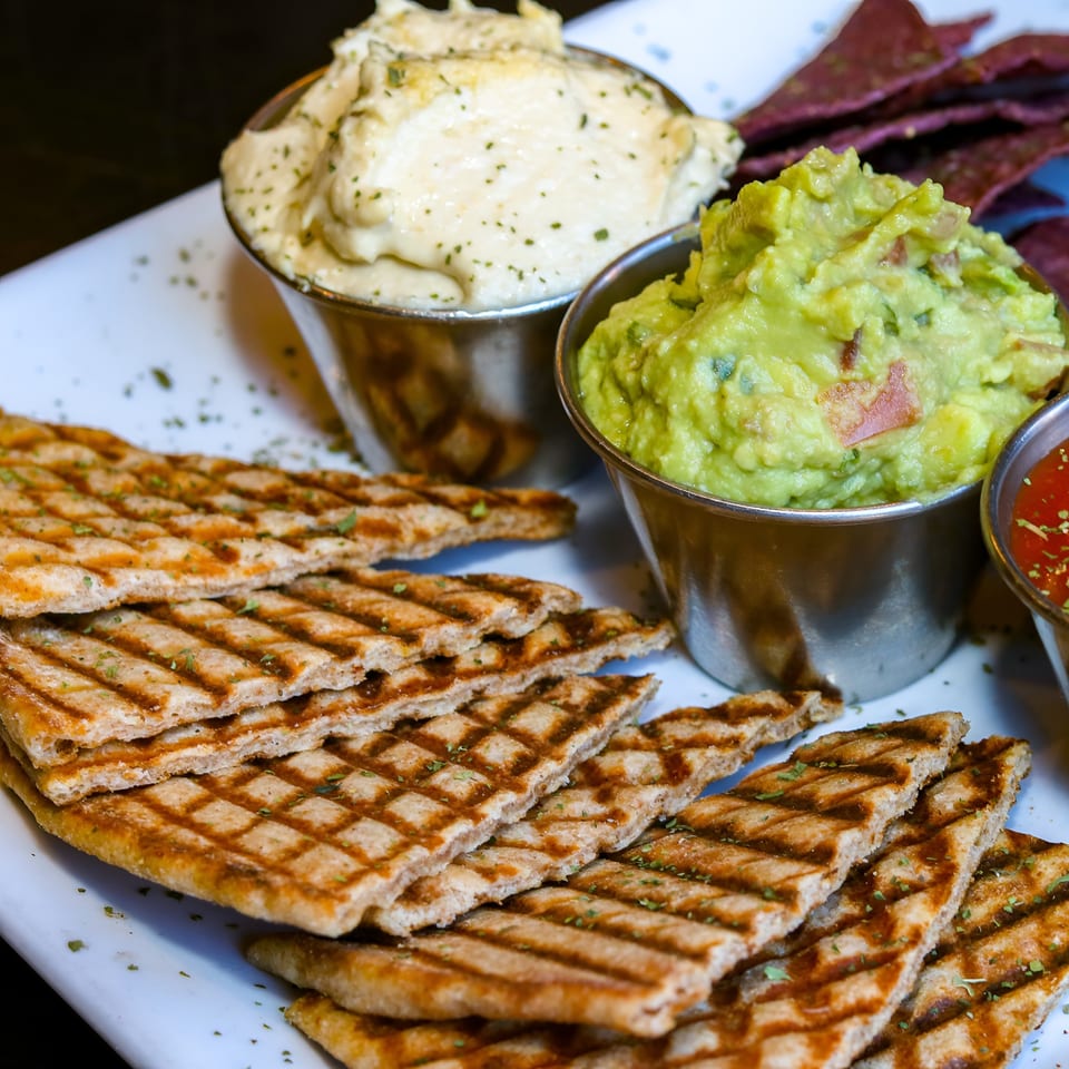A platter with grilled flatbread, hummus, guacamole, salsa, and purple tortilla chips.