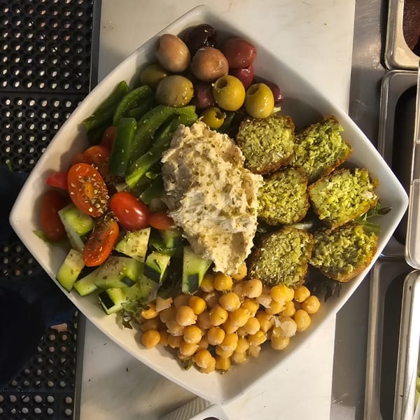 A white square bowl filled with falafel, hummus, chickpeas, olives, tomatoes, cucumbers, and green peppers.