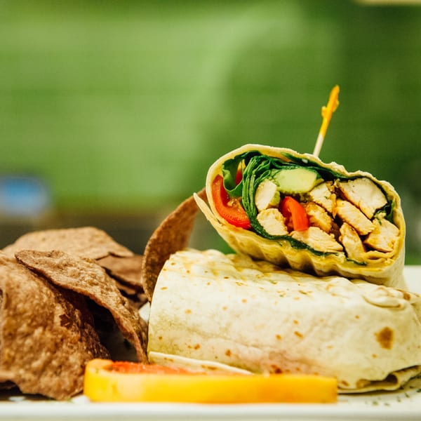 Chicken wrap with lettuce, tomatoes, and cucumbers, served with tortilla chips and a fruit slice.