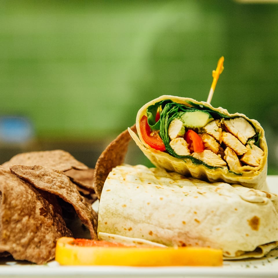 Chicken wrap with lettuce, tomatoes, and cucumbers, served with tortilla chips and a fruit slice.