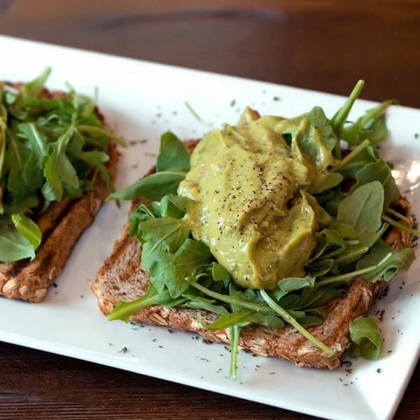 Two slices of avocado toast with arugula and black pepper on a white rectangular plate.