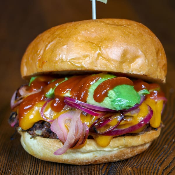 Gourmet burger with melted cheese, red onions, avocado, and sauce on a wooden table.